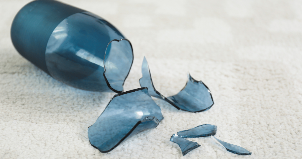 A broken blue glass bottle with shattered pieces scattered on a carpet as shown in Maggy Maid's guide on how to clean broken glass in quick and simple steps.