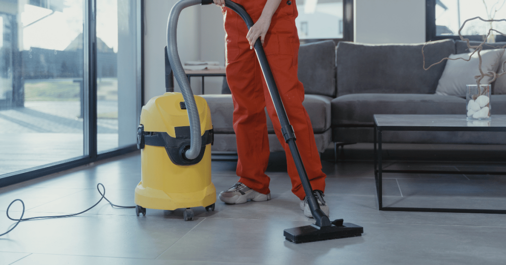 A cleaner in an orange uniform vacuuming a living room floor with an industrial vacuum as shown in Maggy Maid's guide on how to clean broken glass in quick and simple steps.