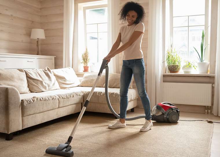 Maggy Maid house cleaning service with a smiling woman vacuuming a carpet in a bright, cozy living room with a canister vacuum