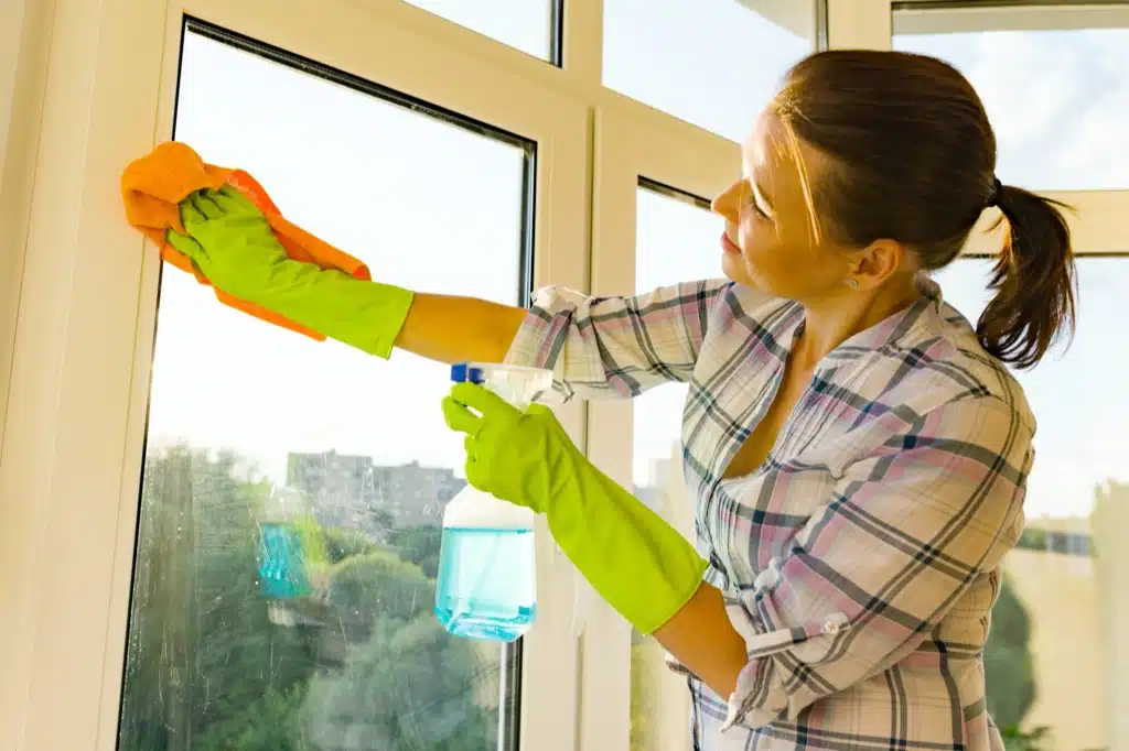 Maggy Maid house cleaner wearing green rubber gloves wiping a window with an orange cloth and blue spray bottle