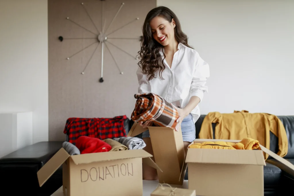 Maggy Maid home decluttering tip with a smiling woman sorting and packing clothes into cardboard donation boxes in a living room