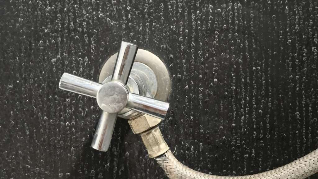 Maggy Maid bathroom cleaning tip showing water stains and mineral deposits on a dark tile wall around a chrome cross-handle shower valve and hose