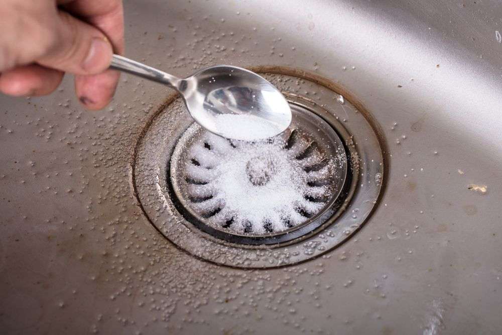 Maggy Maid natural drain cleaning tip with a hand pouring baking soda into a stainless steel kitchen sink drain