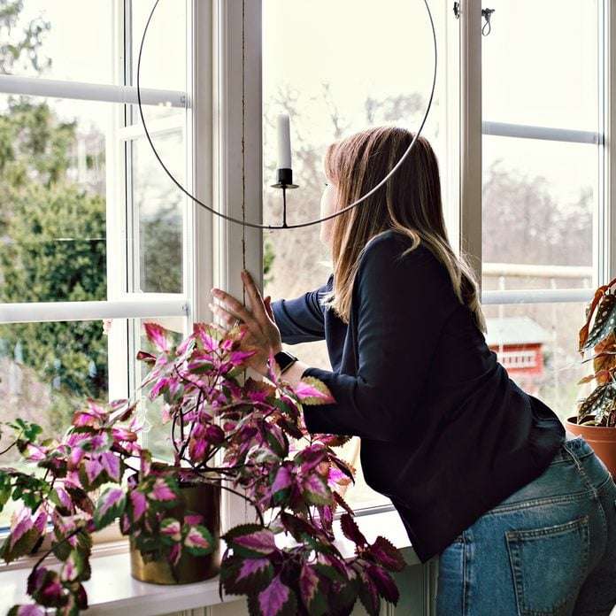 Maggy Maid home cleaning service with a woman wiping a white window frame surrounded by colorful indoor plants on a windowsill